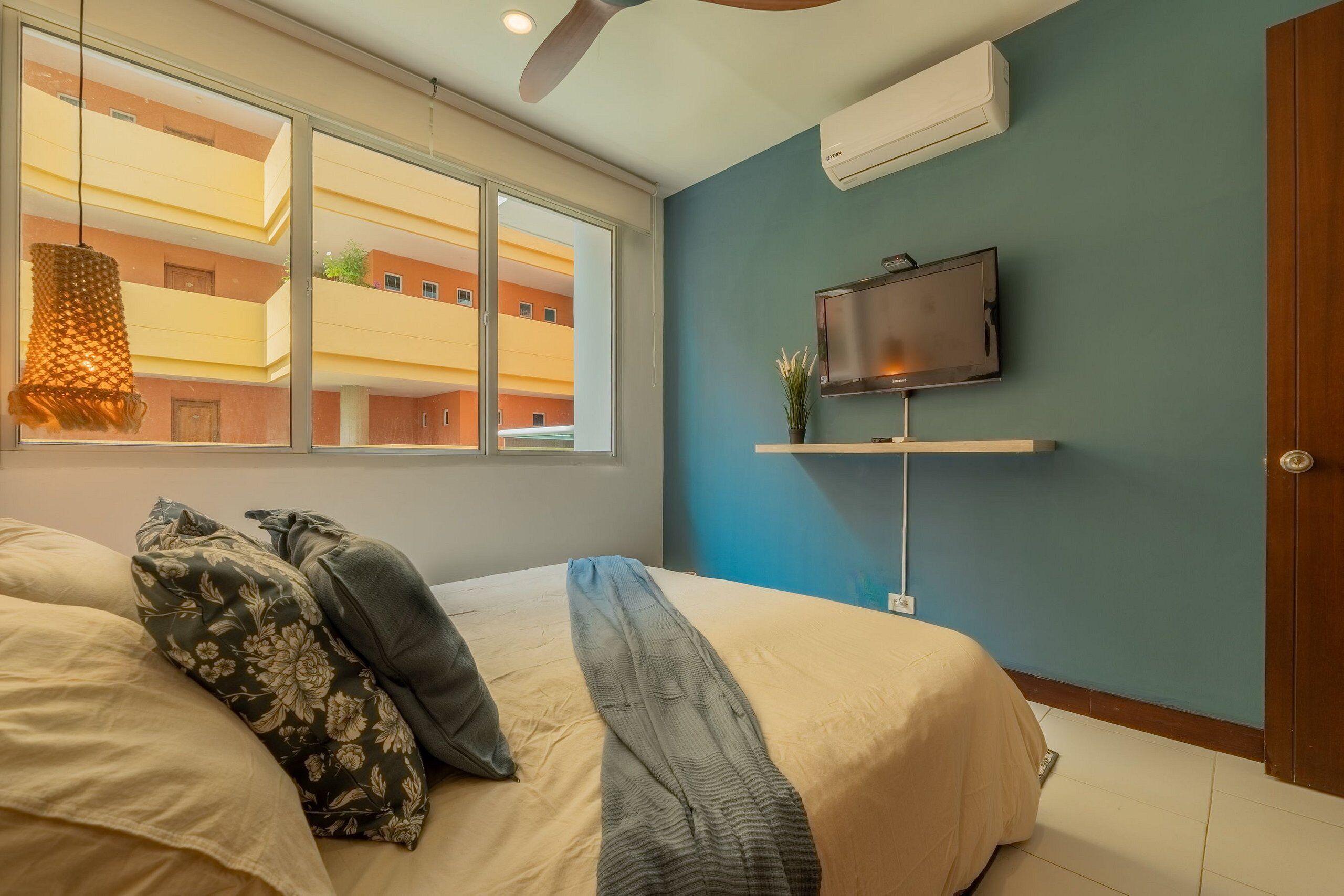 A bedroom with a bed, a TV on a stand, a window, and an air conditioning unit.