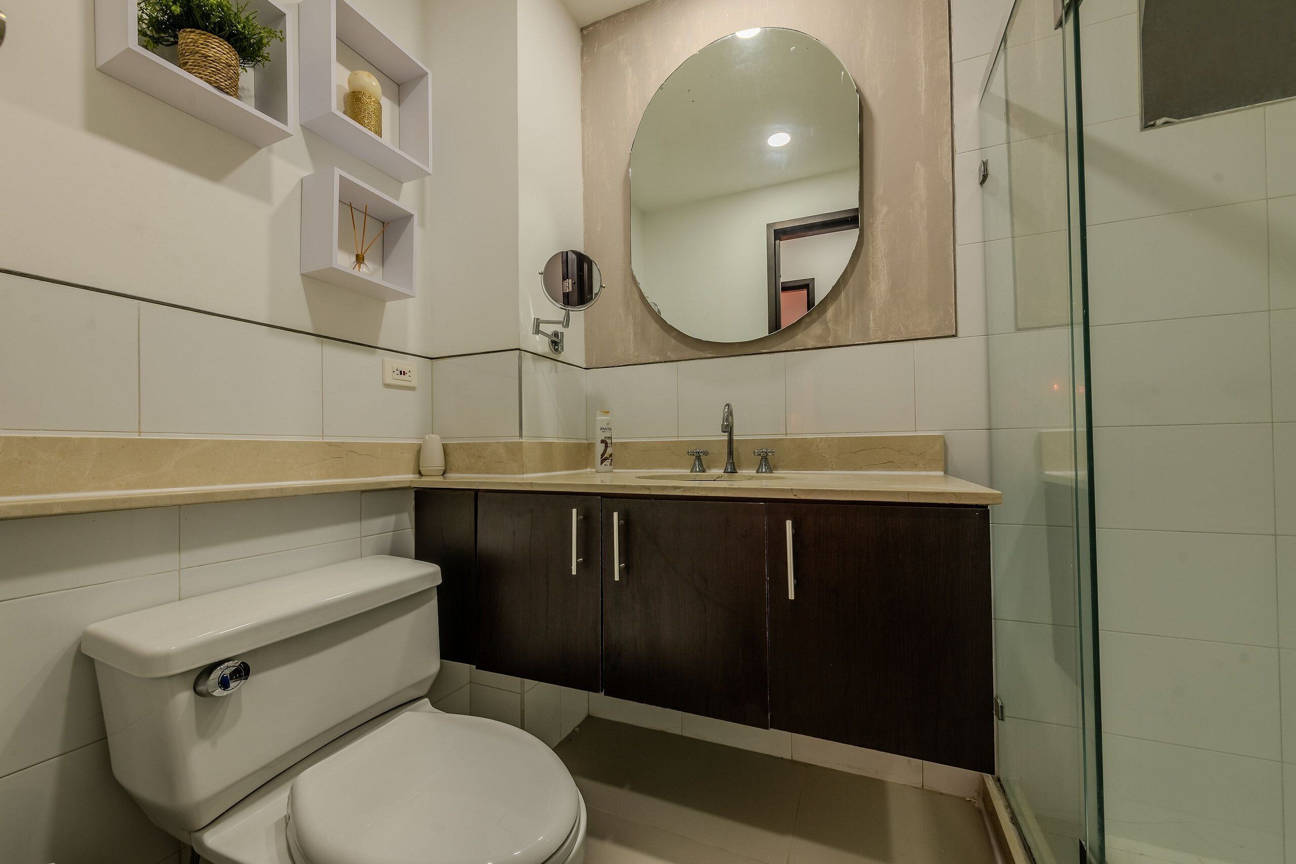 Un baño moderno con inodoro, armarios de madera oscura, un espejo redondo y un box de ducha de vidrio.