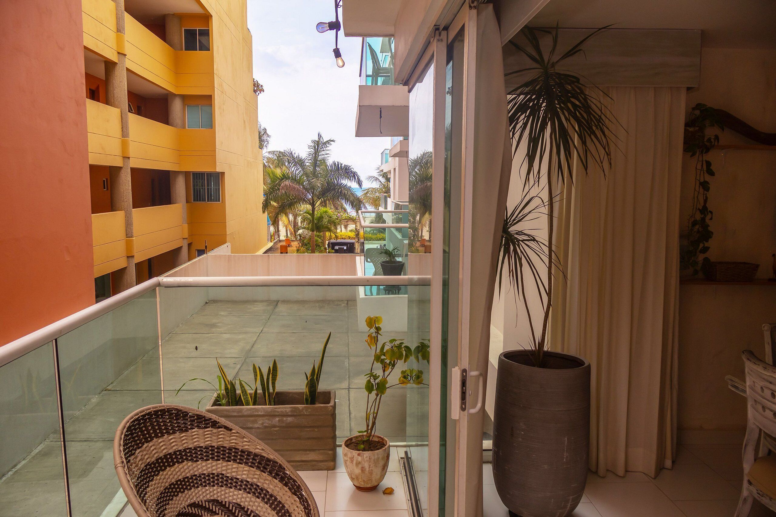 A balcony with a wicker chair, potted plants, and a view of a yellow building and palm trees.