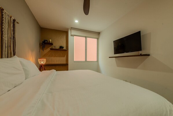 A bedroom with a bed, a TV mounted on the wall, a window with blinds, and a shelf with decorative items.