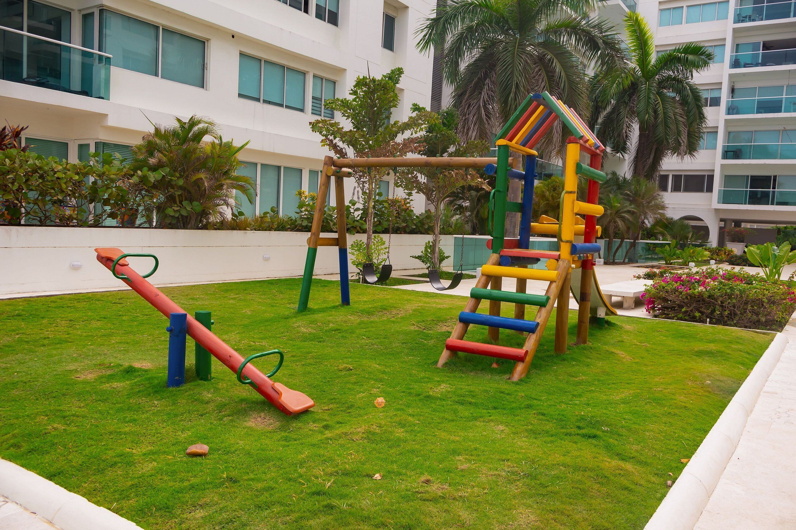 Un parque infantil con tobogán y estructuras para escalar, rodeado de césped cuidado y palmeras, con un edificio al fondo.