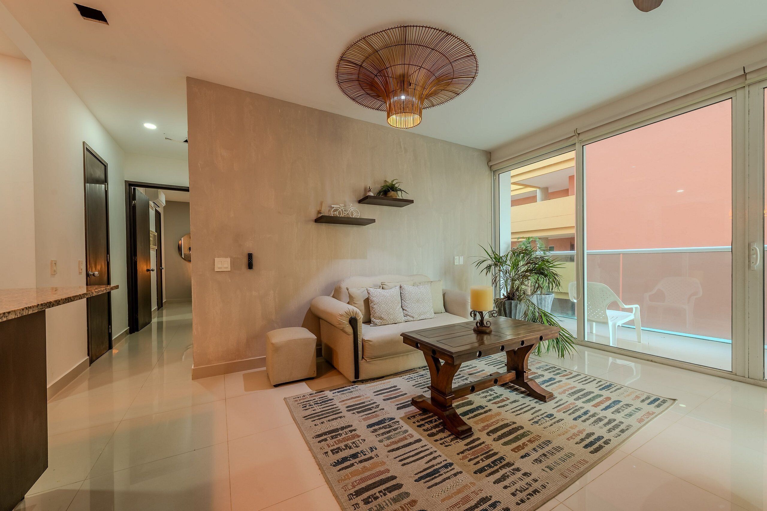 A modern living room with a sofa, a coffee table, and a patterned rug. There is a ceiling light fixture and a sliding glass door leading to an outdoor area.