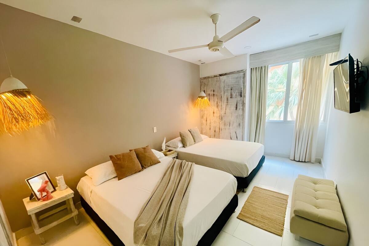 Quarto de hotel com duas camas, um ventilador de teto, televisão e janela com cortinas.