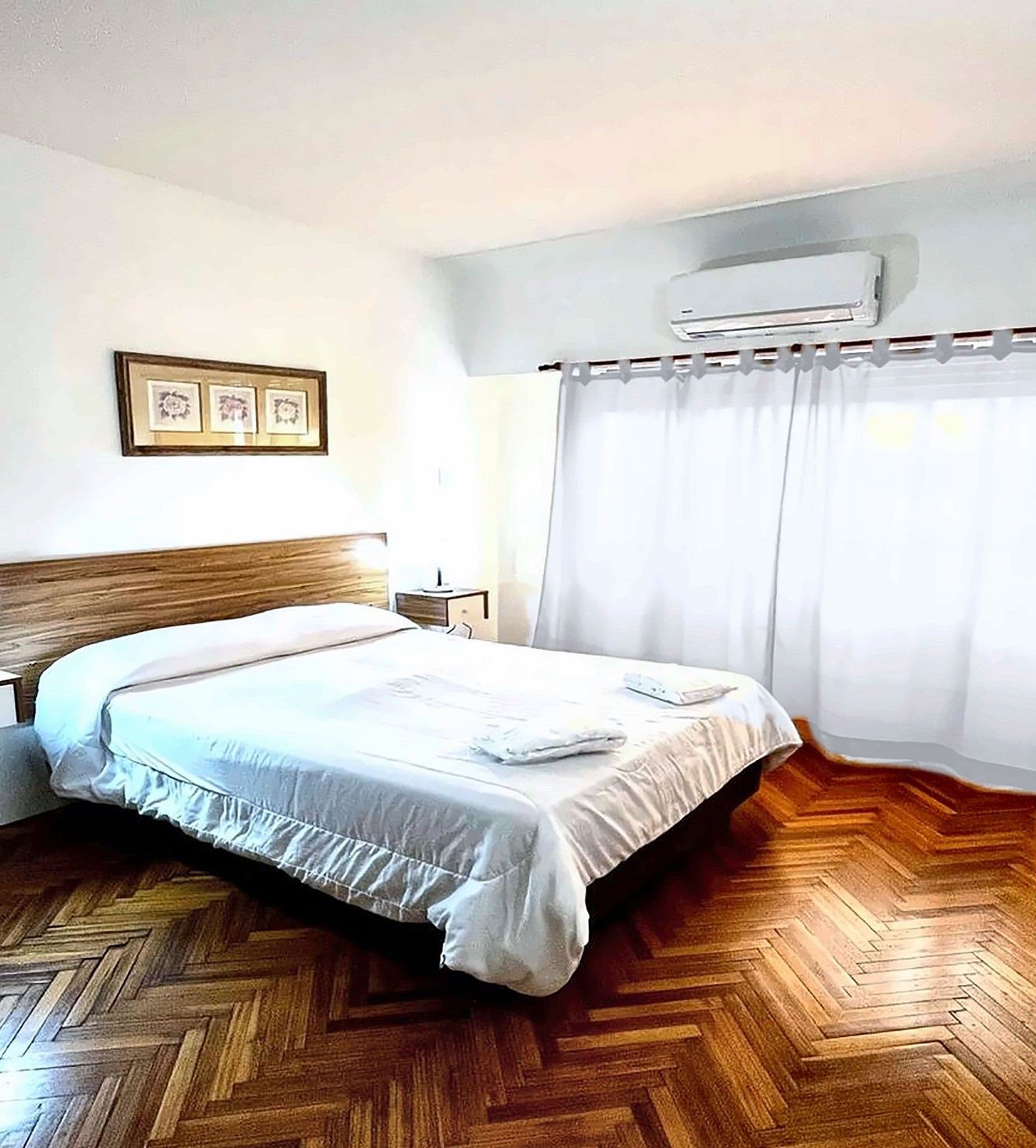 Hotelkamer met een houten visgraatvloer, een bed met witte lakens, een nachtkastje, een ingelijst beeld op de muur en een airconditioner boven het raam.