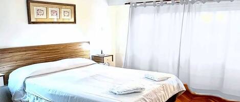 A hotel room with a wooden herringbone floor, a bed with white linens, a nightstand, a framed picture on the wall, and an air conditioning unit above the window.