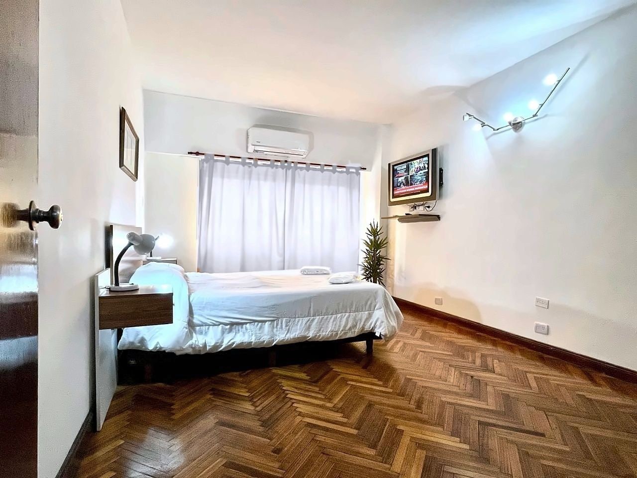 A hotel room with a bed, bedside table, lamp, air conditioning unit, and a television mounted on the wall.