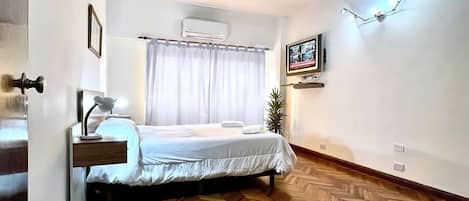 A hotel room with a bed, bedside table, lamp, air conditioning unit, and a television mounted on the wall.