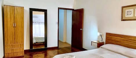 Habitación de hotel con armario de madera, cama con colchón blanco, mesita de noche con lámpara y espejo en la pared.