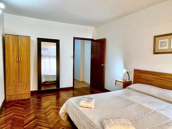A hotel room with a wooden wardrobe, a bed with a white mattress, a bedside table with a lamp, and a mirror on the wall.