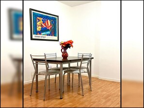Een eethoek met een tafel en vier stoelen, een vaas met bloemen, en een ingelijst schilderij aan de muur.