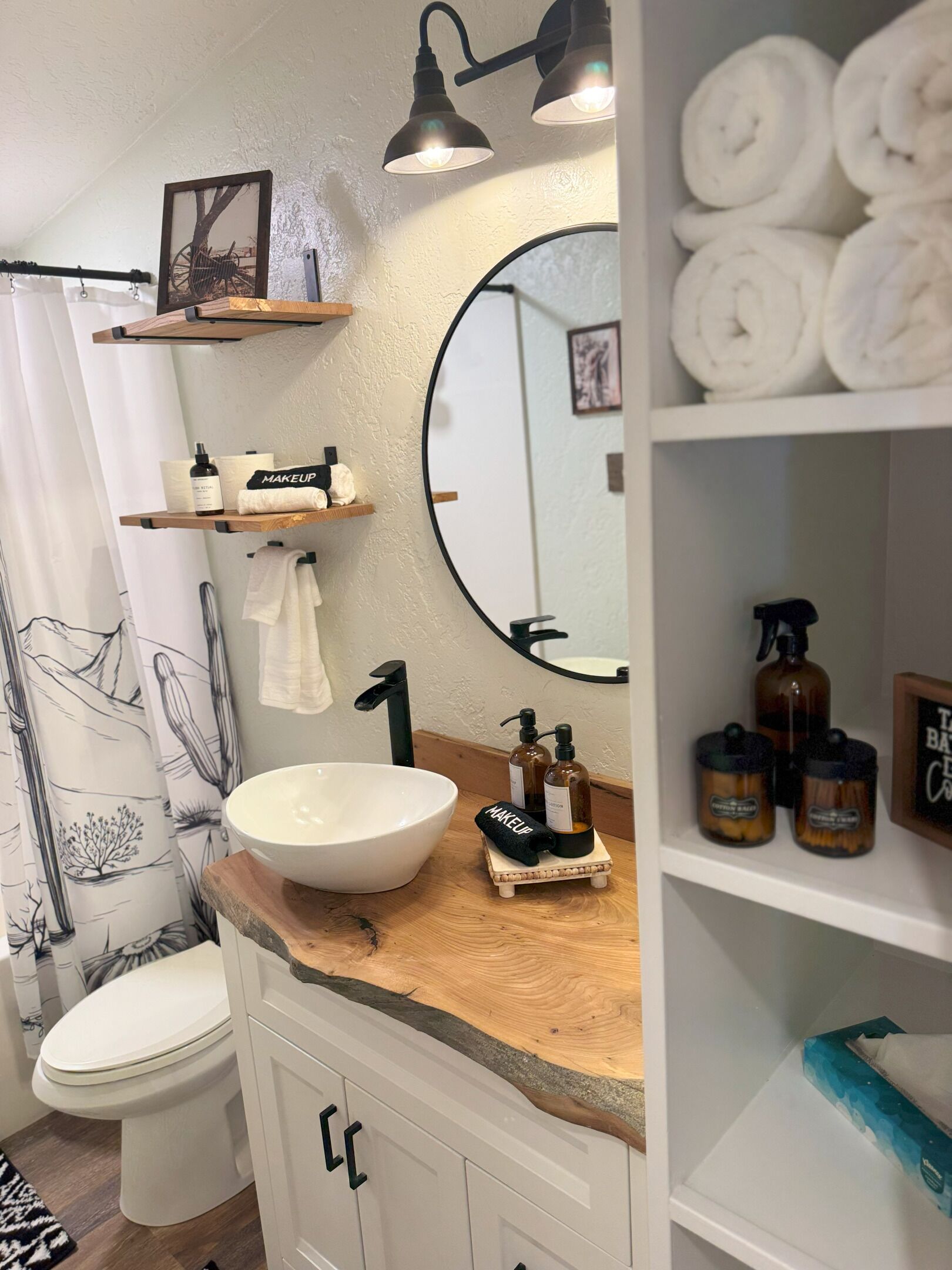 Een badkamer met een wit wastafel, een zwarte kraan en een ronde spiegel. Er zijn planken met handdoeken en toiletartikelen, en een douchegordijn met een zwart-wit ontwerp.