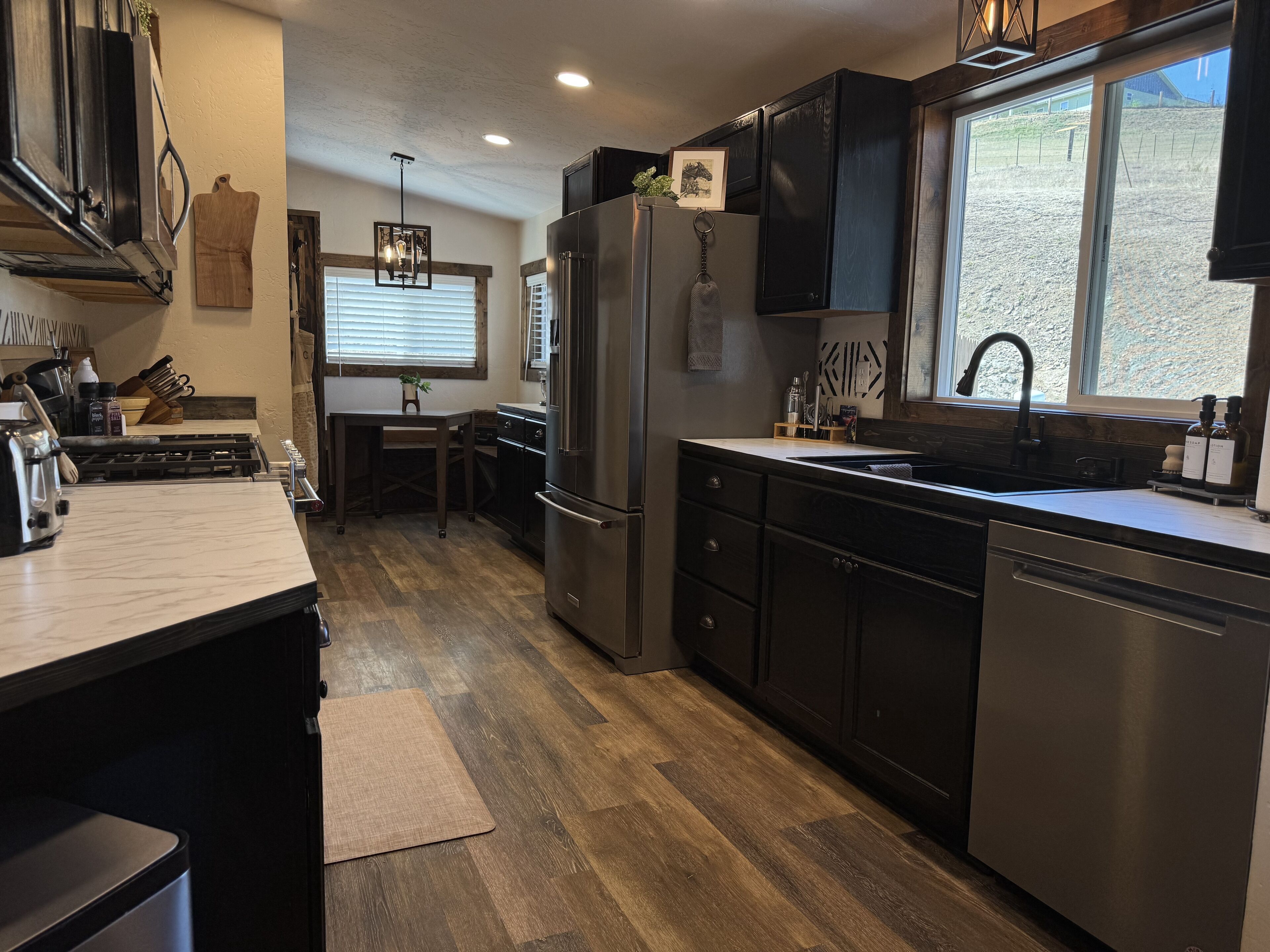 A modern kitchen with dark cabinetry, stainless steel appliances, a wooden dining table, and a large window providing a view of the outdoors.