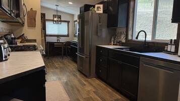 A modern kitchen with dark cabinetry, stainless steel appliances, a wooden dining table, and a large window providing a view of the outdoors.