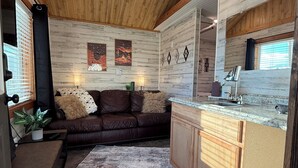 A compact living space with a wooden ceiling, a sofa, a small table with a plant, a kitchen area with a sink and cabinets, and a rug on the floor. - 4BR Ranch Retreat w/Hot Tub & Senic Valley Views + Mini House Add-On (Kamiah)