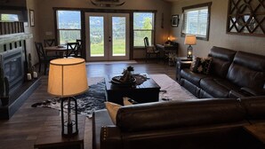 A spacious living room with a fireplace, leather sofa, and a large window offering a view of the outdoors. - 4BR Ranch Retreat w/Hot Tub & Senic Valley Views + Mini House Add-On (Kamiah)