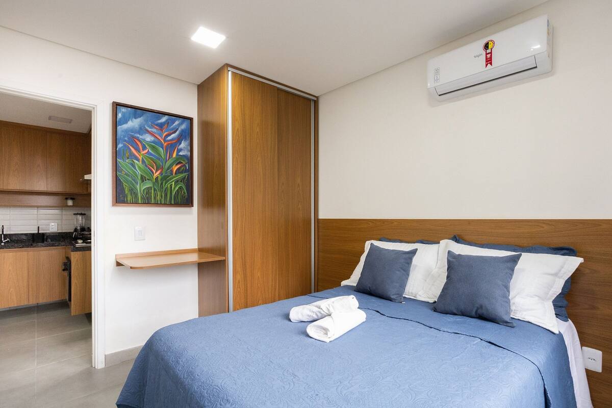 A hotel room with a bed, a painting, a kitchen area, and an air conditioning unit.