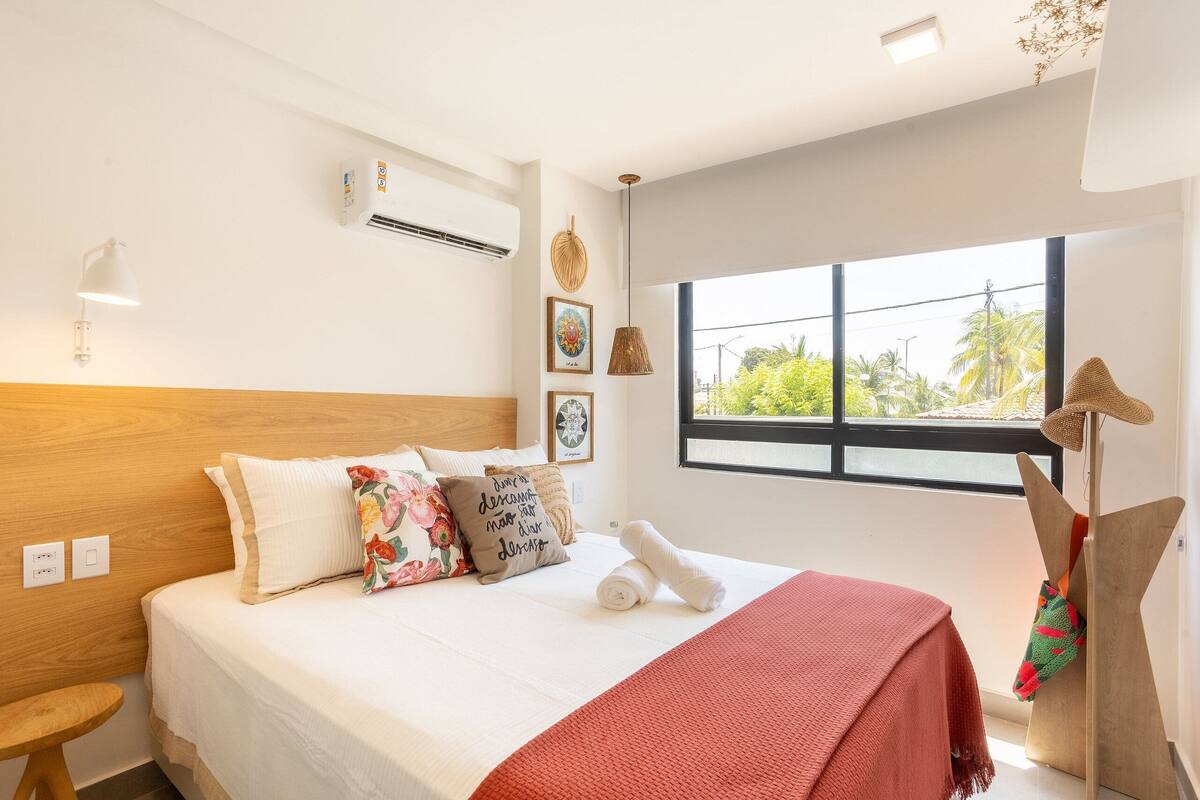A neatly made bed with pillows and a blanket, a bedside table, a window with a view, and an air conditioning unit on the wall.