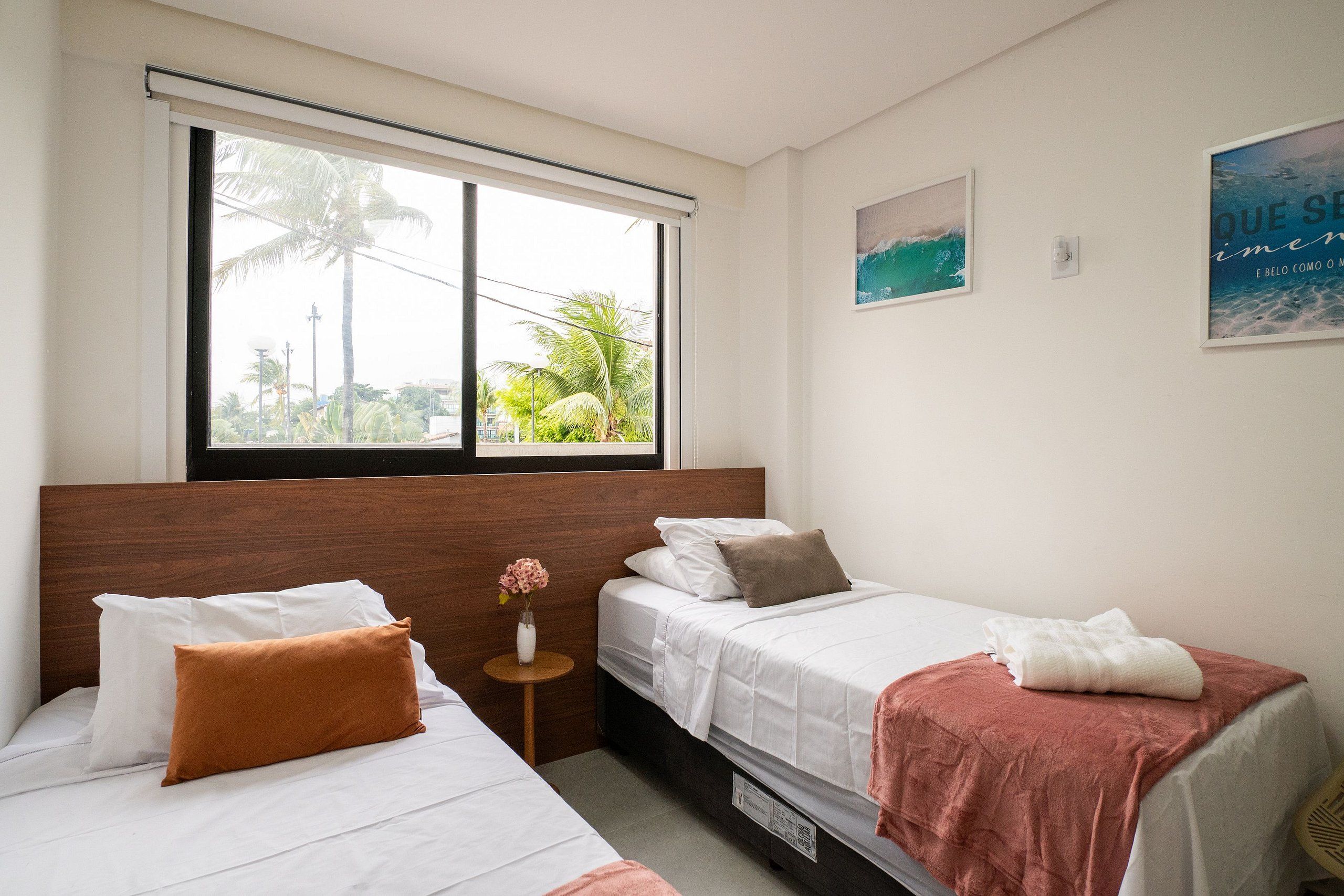 A hotel room with two beds, a window with a view of palm trees, and a small table with a vase.