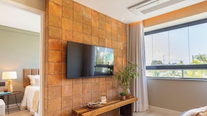 Une chambre d’hôtel moderne dotée d’un téléviseur à écran plat fixé à un mur en pierre, d’un banc en bois, d’une table de chevet avec une lampe et d’une grande fenêtre offrant une vue sur les palmiers.