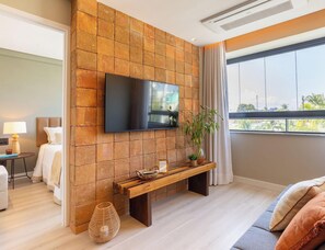 Una habitación de hotel moderna con un televisor de pantalla plana montado en una pared de piedra, un banco de madera, una mesita de noche con lámpara y un gran ventanal con vistas a palmeras.