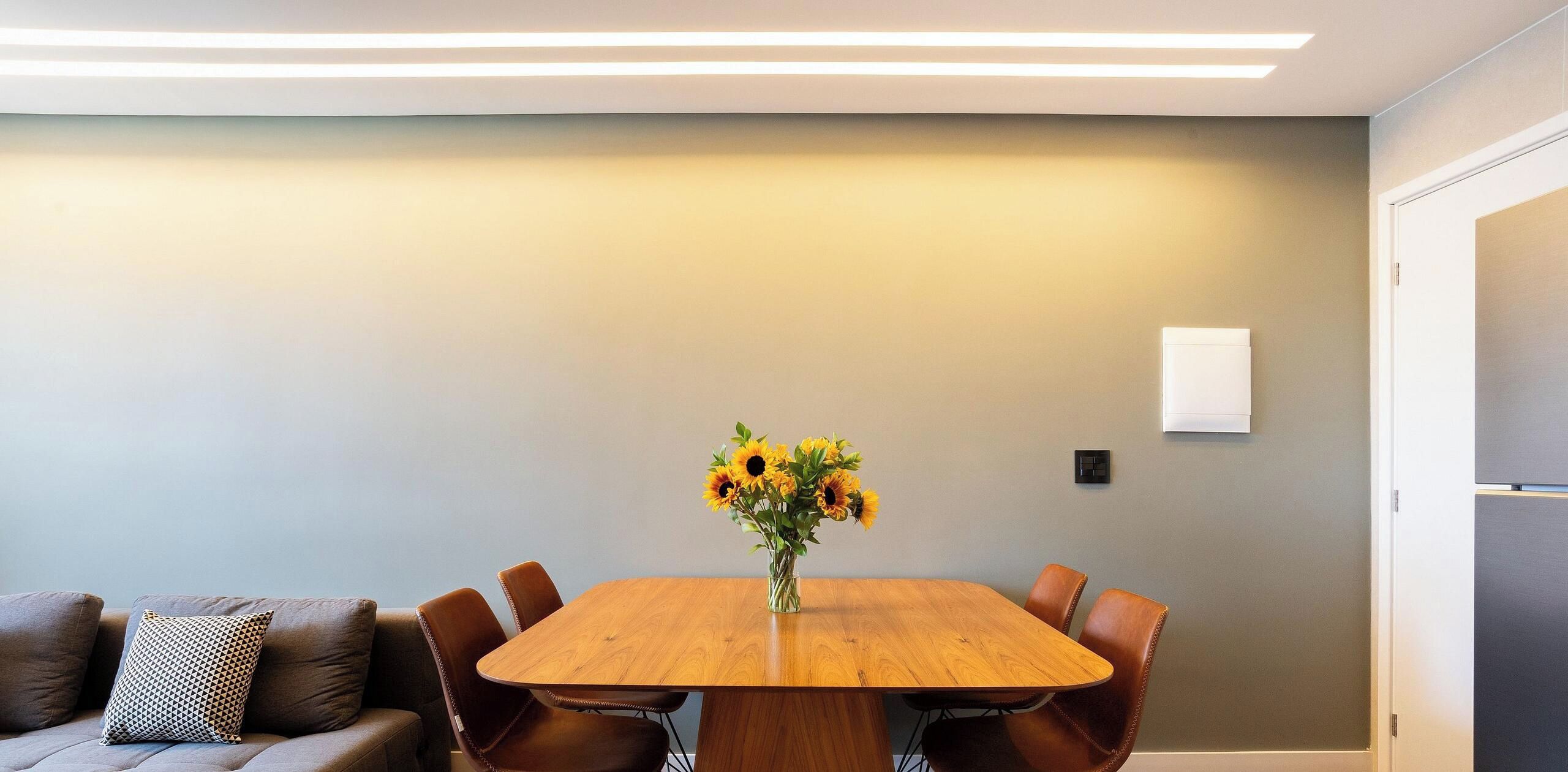 A dining area with a wooden table, six chairs, a vase with sunflowers, a sofa with patterned cushions, and a wall-mounted light fixture.