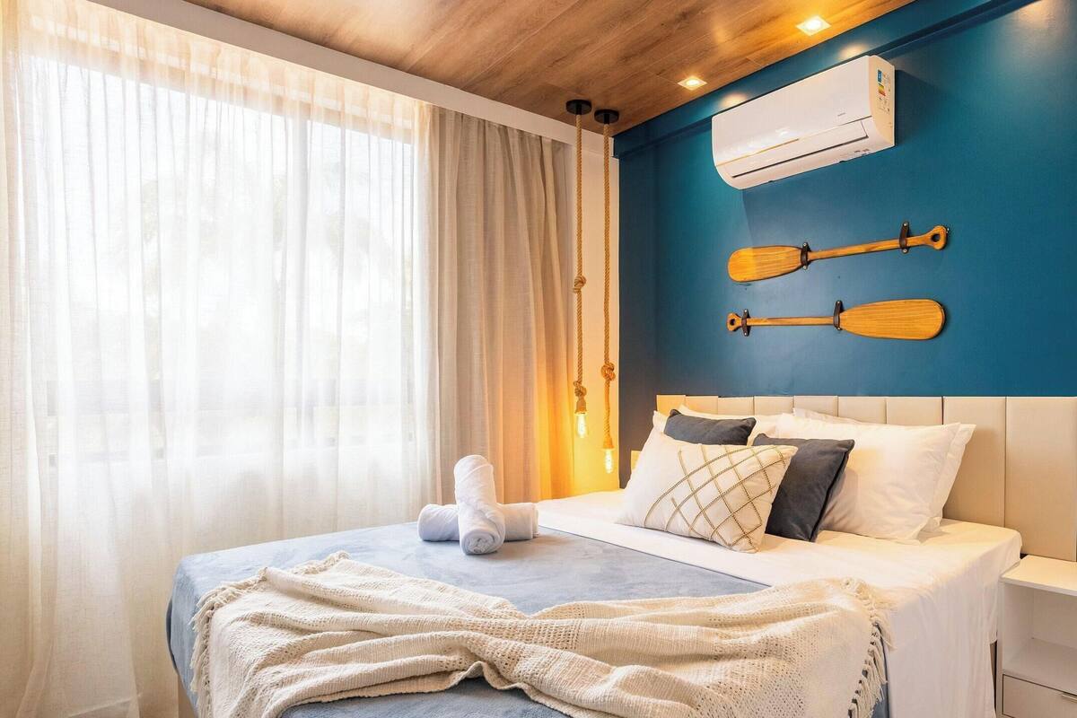 A bedroom with a bed, white bedding, a blue wall, an air conditioning unit, and two wooden paddles.
