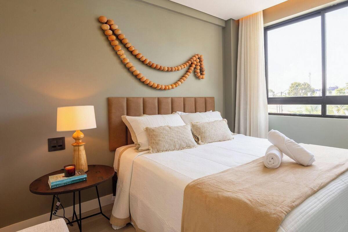 A hotel room with a bed, a bedside table with a lamp, a wall art piece, and a window with curtains.