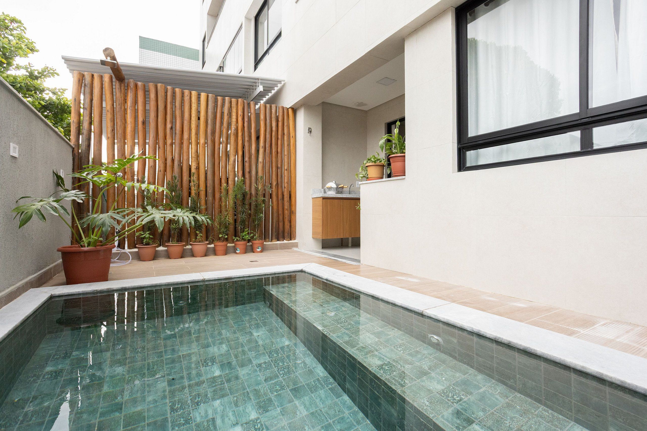 Une piscine dont les bords sont recouverts de carreaux, agrémentée de plantes en pot, entourée d’une clôture en bois et dotée d’une fenêtre avec un rideau.