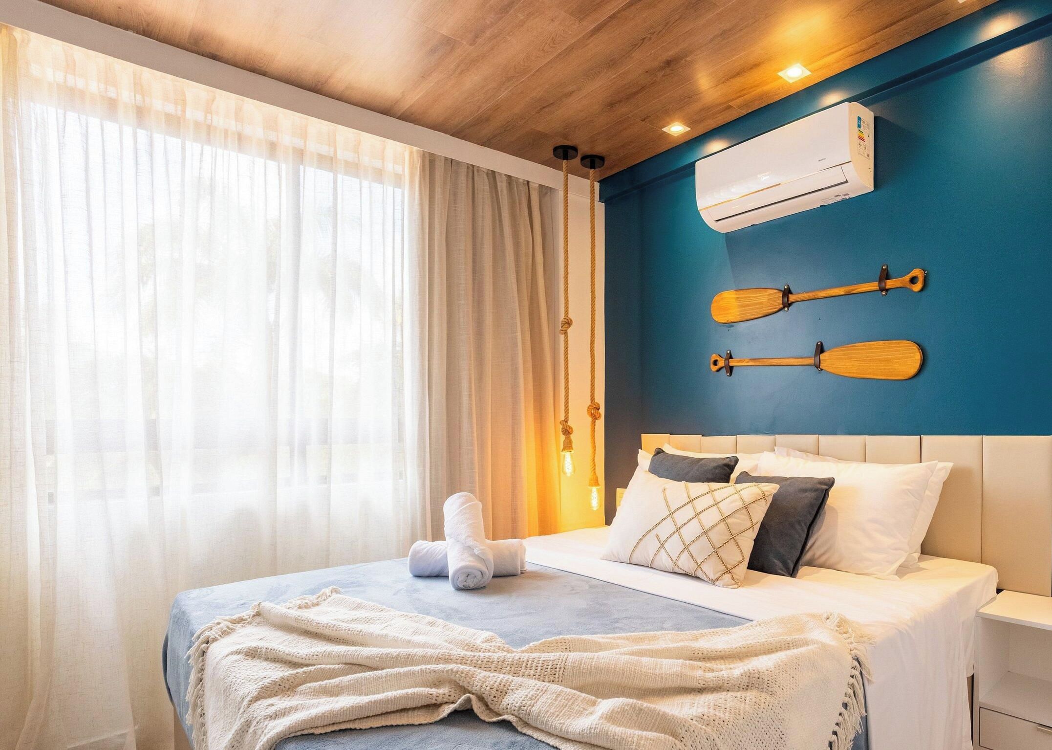 Een hotelkamer met een bed, een raam met gordijnen, een airconditioning en twee houten spatborden aan de muur.