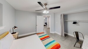 A hotel room with a bed, a desk, a chair, a ceiling fan, and a door leading to another room.