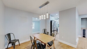 A dining area with a wooden table and four chairs, a hanging light fixture, and a kitchen in the background.