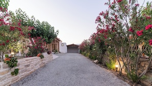 A gravel driveway leading to a house with a stone wall on one side and a wooden fence on the other, surrounded by flowering plants.