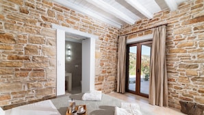 A stone-walled room with a bed, a tray with cups and a teapot, and a view through a glass door.