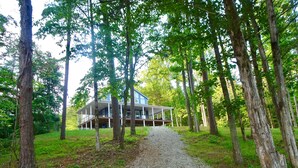 A house with a covered porch, surrounded by tall trees and a gravel path leading to it. - Forget Me Not Haven – 6BR Lakefront Home for Unforgettable Family Memories (Speedwell)