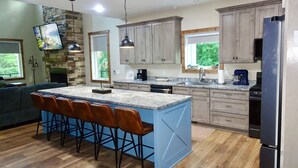 A modern kitchen with a large island, bar stools, wooden cabinets, and a stone accent wall. - Forget Me Not Haven – 6BR Lakefront Home for Unforgettable Family Memories (Speedwell)