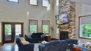 A spacious living room with a stone fireplace, a large TV mounted above it, and a view of the outdoors through multiple windows. - Forget Me Not Haven – 6BR Lakefront Home for Unforgettable Family Memories (Speedwell)
