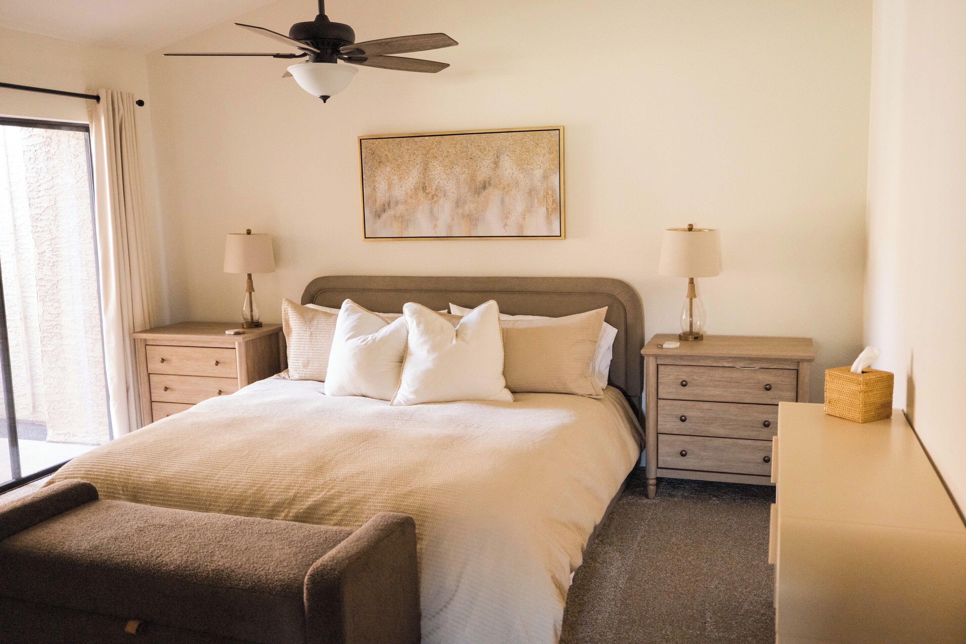 A neatly arranged hotel room with a bed, two bedside tables, a ceiling fan, a wall-mounted painting, and a window with curtains.