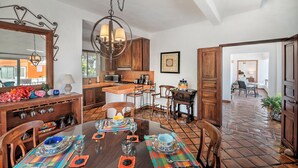 Un comedor con una mesa de madera preparada para cuatro personas, rodeada de sillas. Se ve una cocina con armarios de madera y una lámpara colgante al fondo.