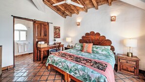 Un dormitorio con techo de madera, una cama con colcha estampada, un escritorio con silla, una mesita de noche con lámpara y una lámpara de pared.