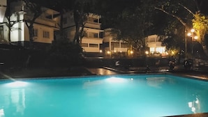 Un espace piscine éclairé pour la baignade nocturne, offrant une vue dégagée sur la piscine, les arbres environnants et les bâtiments adjacents.