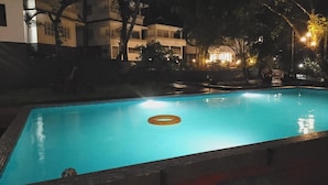 Une piscine éclairée la nuit, avec un grand flotteur gonflable visible, entourée d’arbres et de bâtiments.