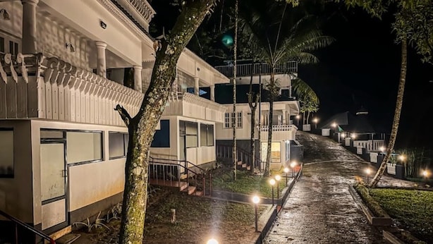 Un chemin mène à une rangée de bâtiments blancs dotés de balcons, entourés d’arbres et éclairés par des lampadaires.