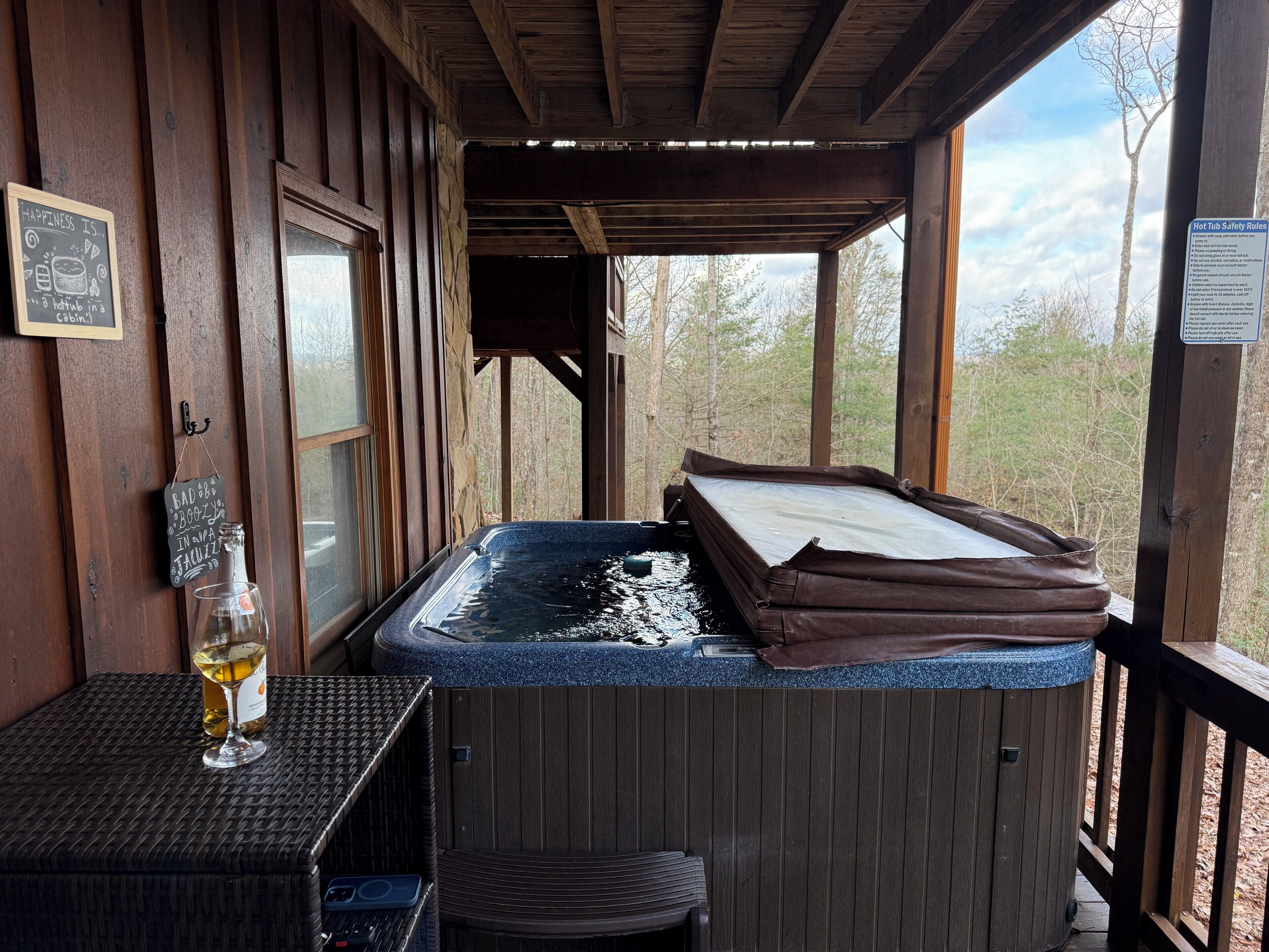 Un bain à remous sur une terrasse en bois, avec vue sur les arbres, un verre de vin et un panneau indiquant les règles de sécurité.