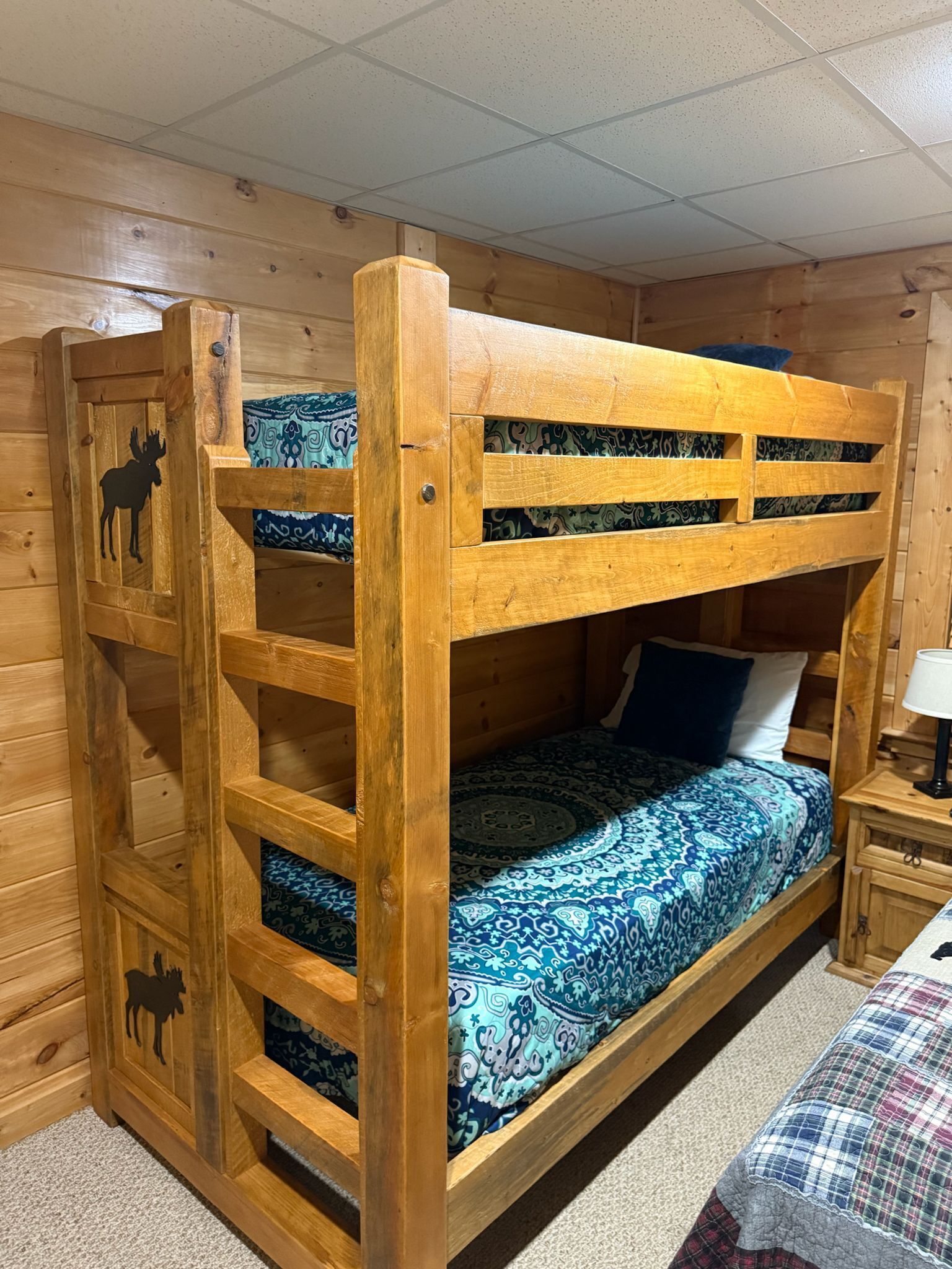 Un lit superposé en bois avec une échelle, une literie à motifs et une petite table de chevet avec une lampe.