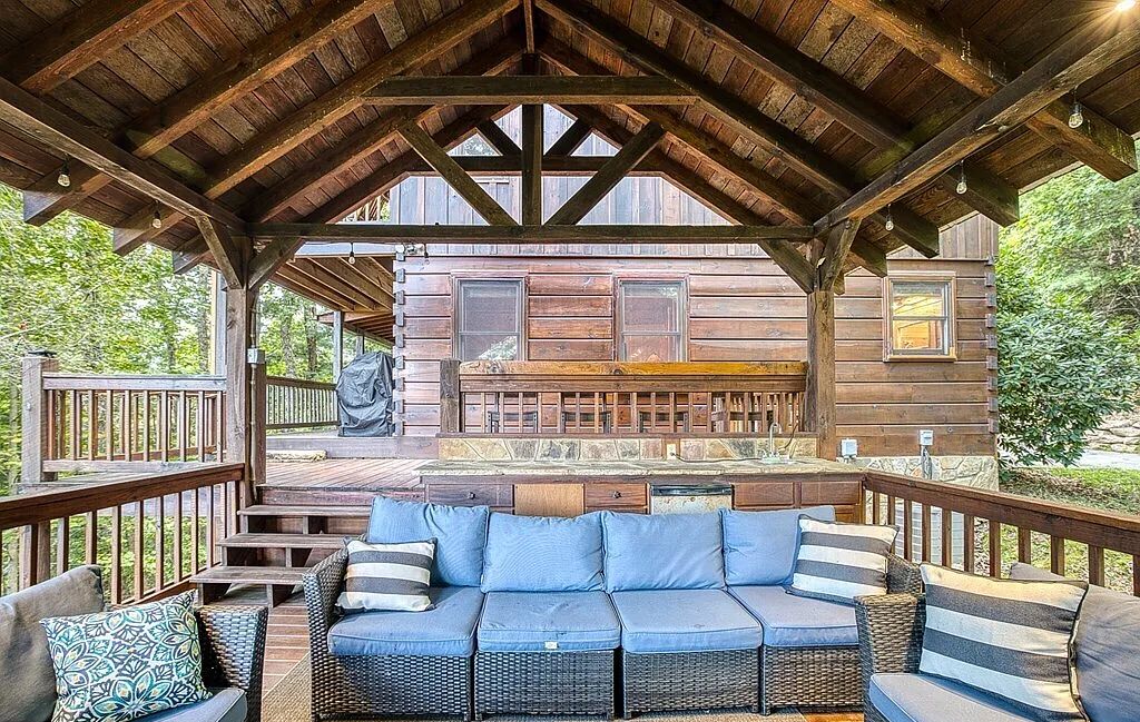 Ein überdachter Außenbereich mit Holzdeck, einer Couch mit Kissen und Blick auf eine rustikale Holzhütte.