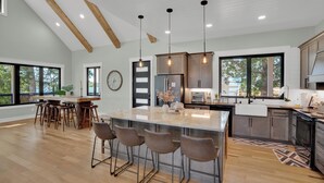 A spacious kitchen with a large island, bar stools, wooden ceiling beams, pendant lights, and large windows. - Luxurious Lakefront retreat with own private dock. (Chapin)