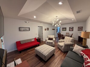 Amplia sala de estar con un sofá rojo, un taburete estampado y sillones, suelo de madera y una zona de comedor con sillas y una mesa.