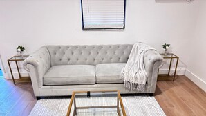 A living room with a tufted grey sofa, a glass coffee table, and two side tables with plants.