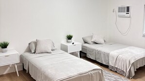 A minimalist bedroom with two single beds, a ceiling fan, an air conditioning unit, and a small bedside table with a plant.