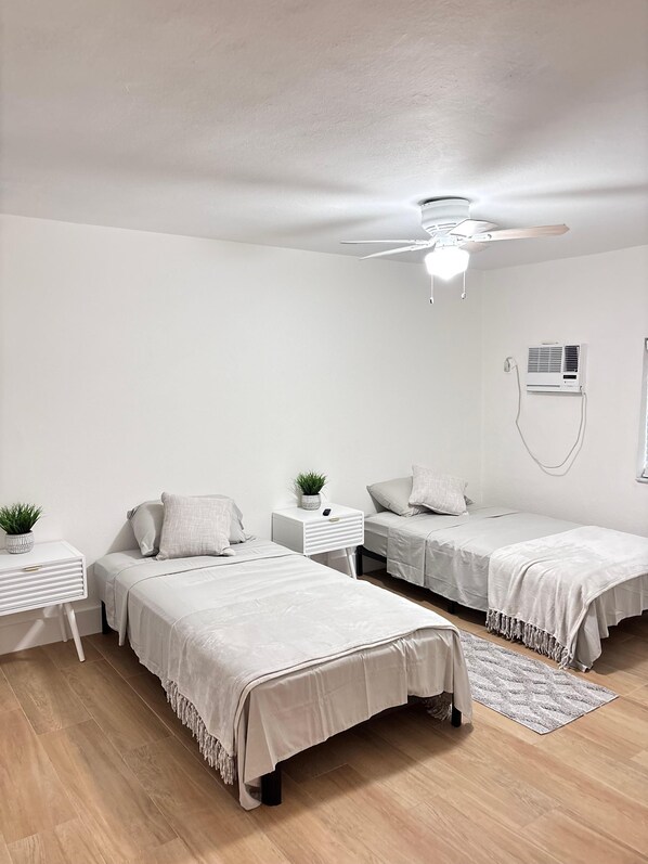 A minimalist bedroom with two single beds, a ceiling fan, an air conditioning unit, and a small bedside table with a plant.